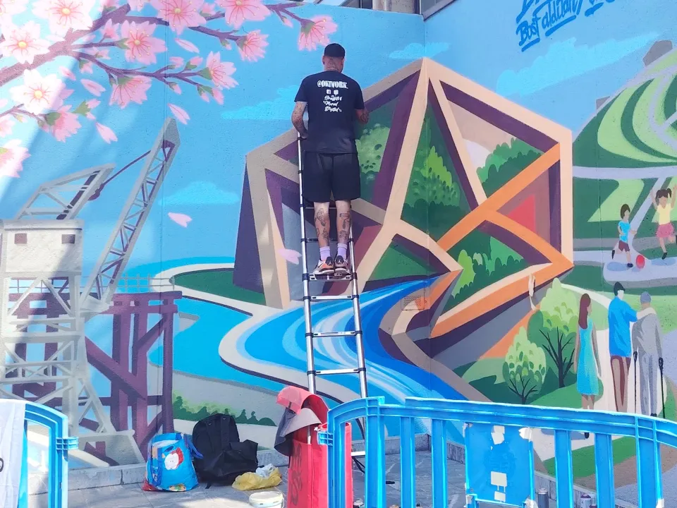 Okiman subido a una escalera pintando el mural de Barakaldo, con el cerezo en flor y la grúa ya terminados