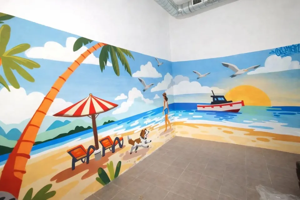 Mural en una esquina interior con una playa, una sombrilla, gaviotas y un barco