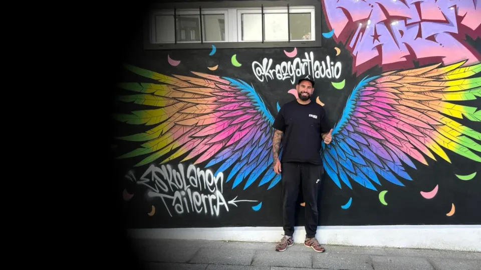 Okiman con el pulgar hacia arriba delante de un mural de alas de colores
