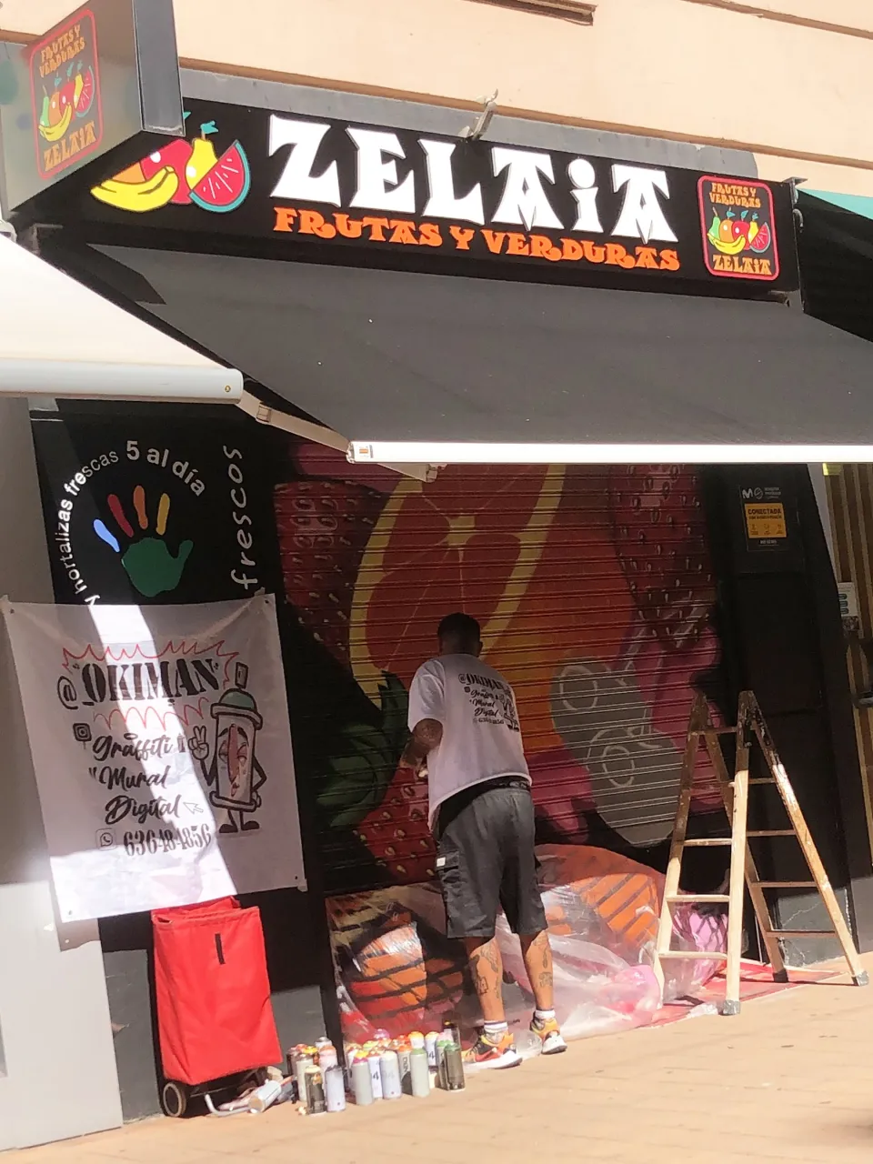 Okiman pintando la persiana de la frutería Zelaia, con los botes de spray en el suelo