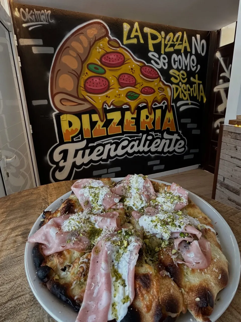 Pared negra de una pizzería con una porción de pizza y el lema «La pizza no se come, se disfruta»